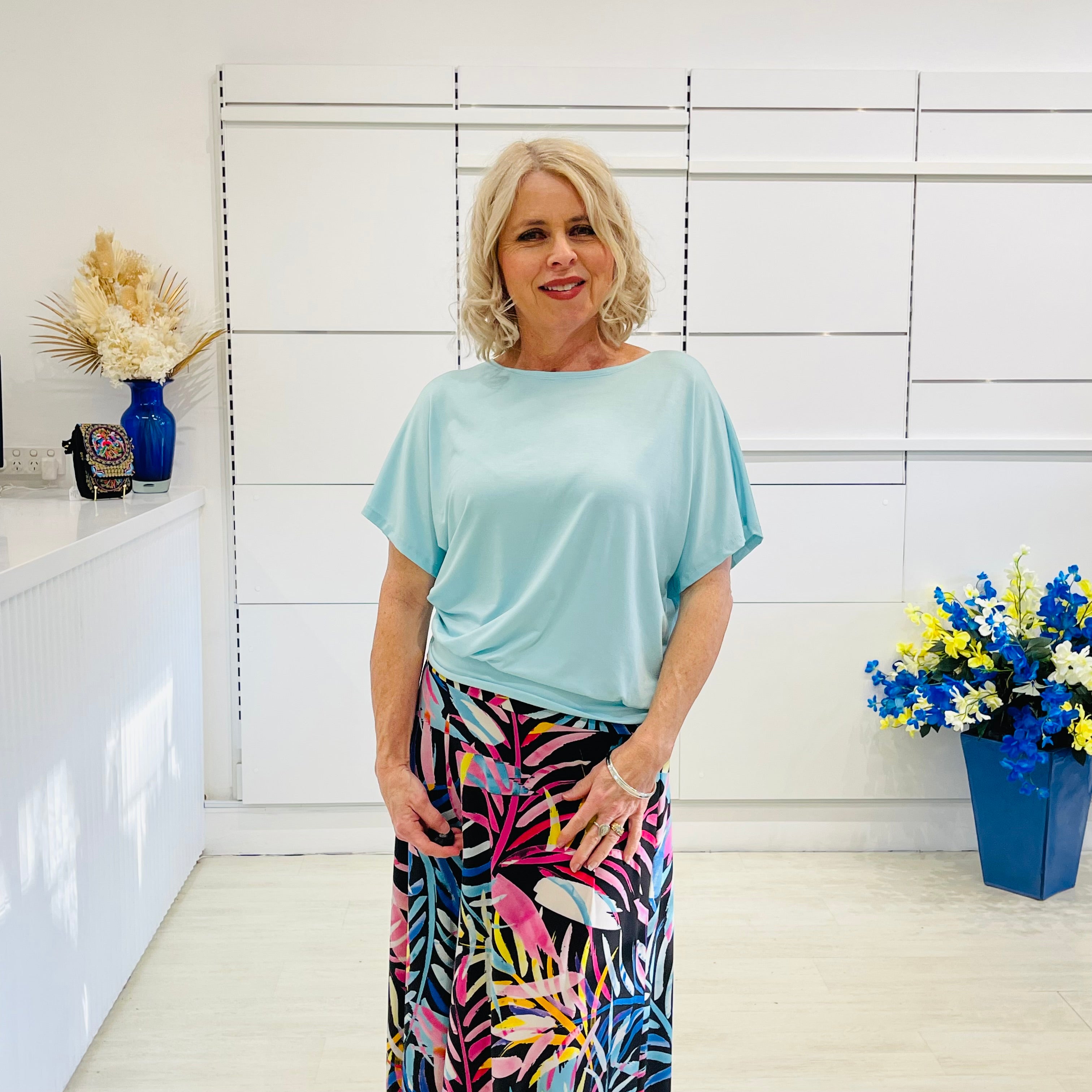 Woman wearing a light blue top and colorful skirt in a room with white walls and decorative elements.