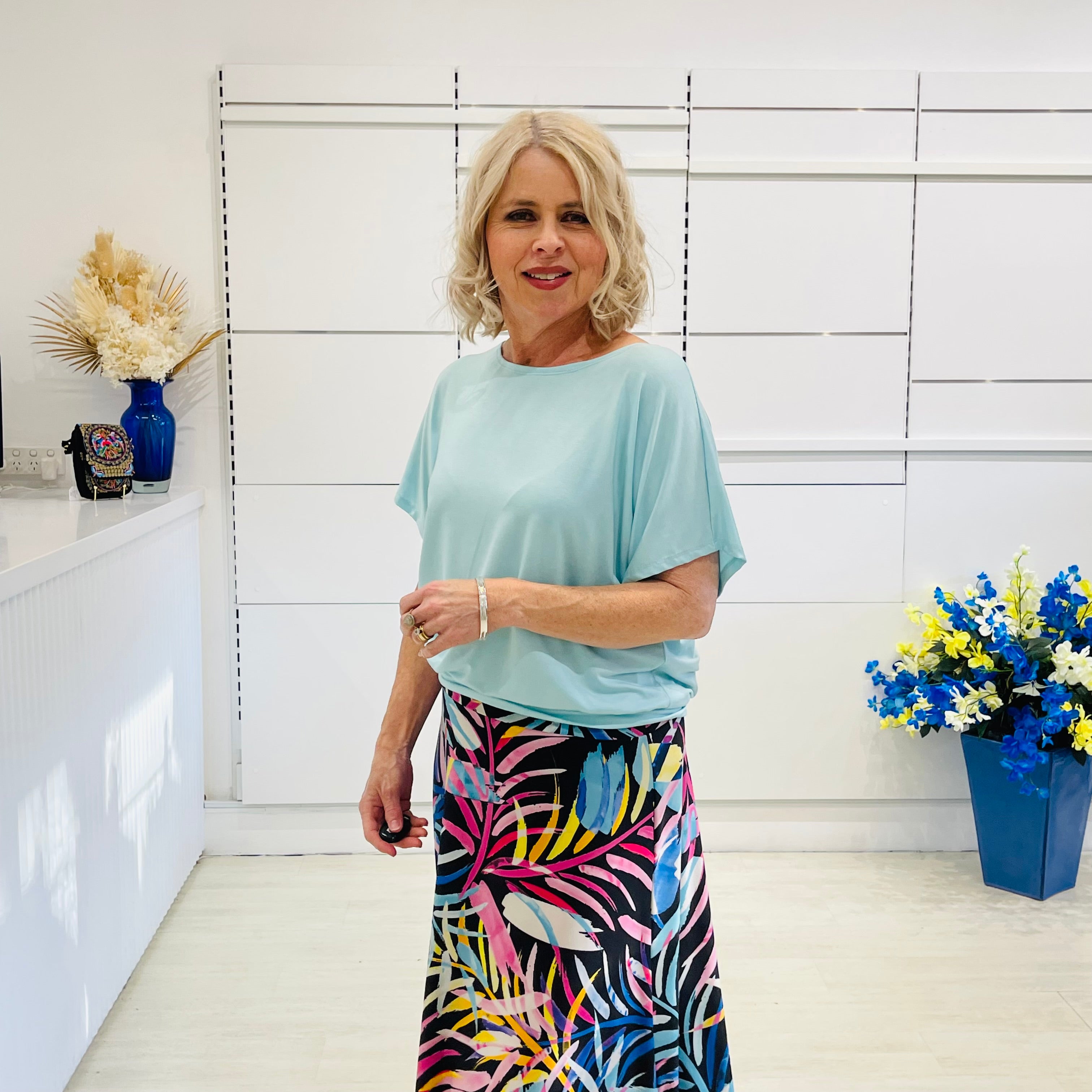Woman wearing a light blue top and colorful skirt in a room with white walls and decorative elements.