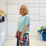 Woman wearing a light blue top and colorful skirt standing in a room with white walls and decorative elements.