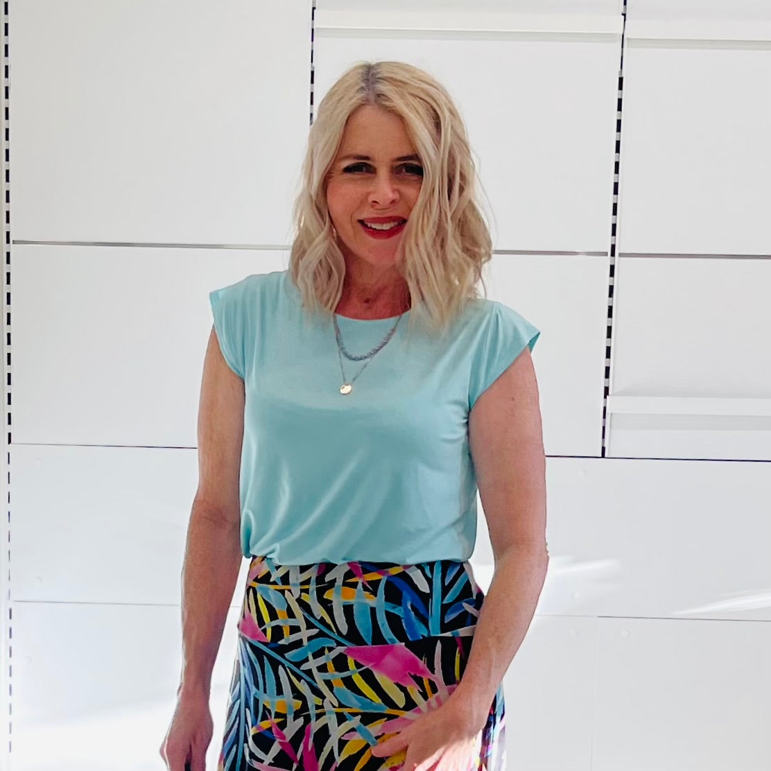 Woman wearing a light green top and colorful skirt against a white tiled wall.