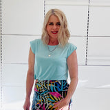 Woman wearing a light green top and colorful skirt against a white tiled wall.
