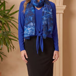 Woman wearing a blue top and black skirt with a blue patterned scarf, standing against a beige wall.