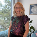 Woman wearing a colorful sequined top indoors with a window and plant in the background