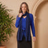 Woman wearing a blue outfit with a scarf, standing next to a potted plant against a beige wall.
