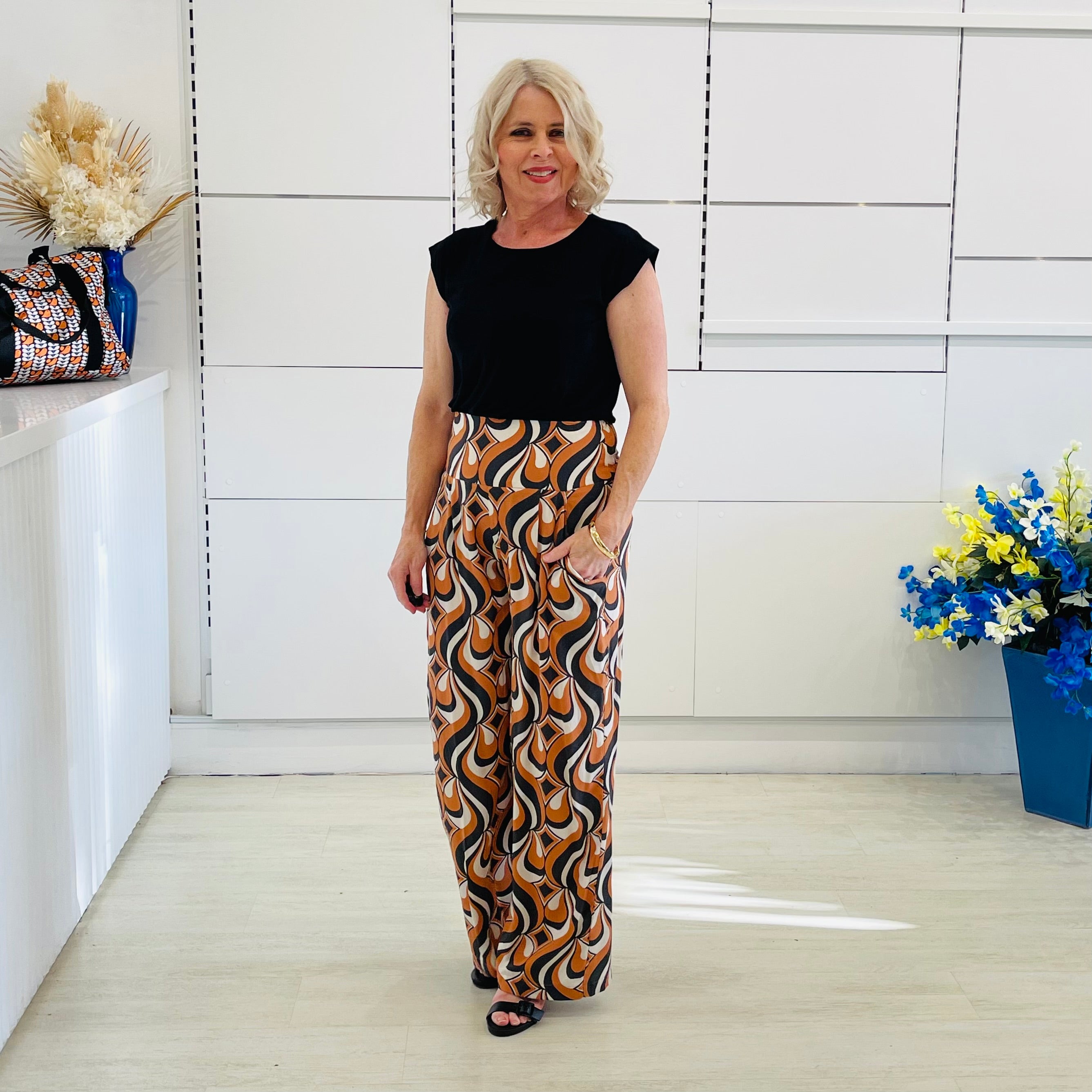 Woman wearing a black top and patterned pants standing in a room with white walls and decorative elements.