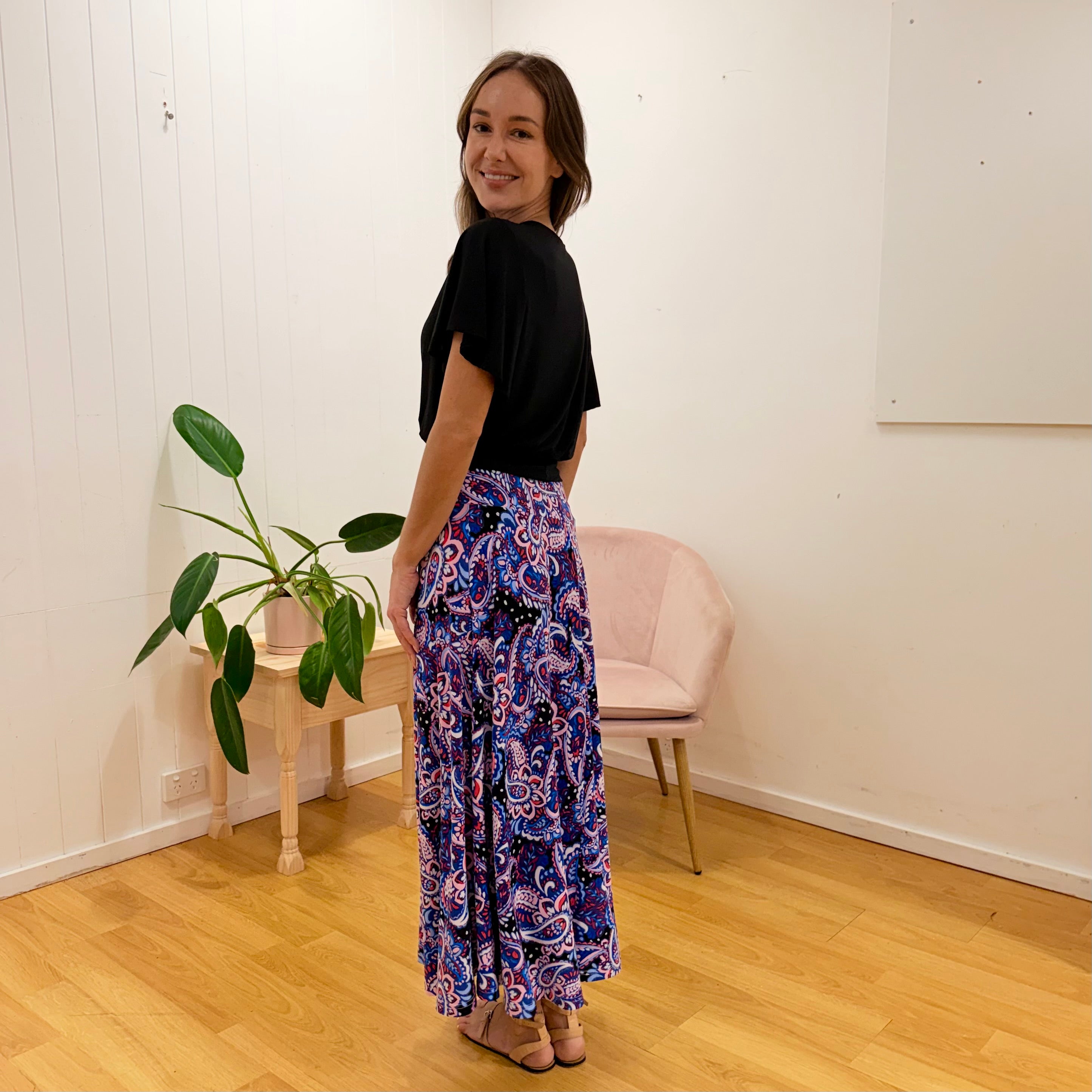 Woman wearing a black top and colorful patterned pants standing in a room with a plant and chair.