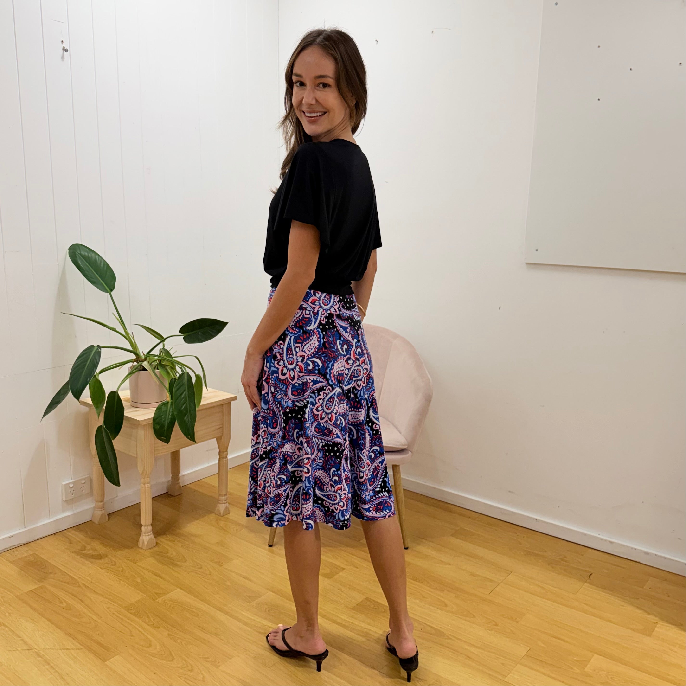 Woman wearing a black top and colorful paisley skirt standing in a room with a plant and chair.