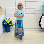 Woman in a light blue top and colorful patterned skirt standing in a room with white walls and a flower arrangement.