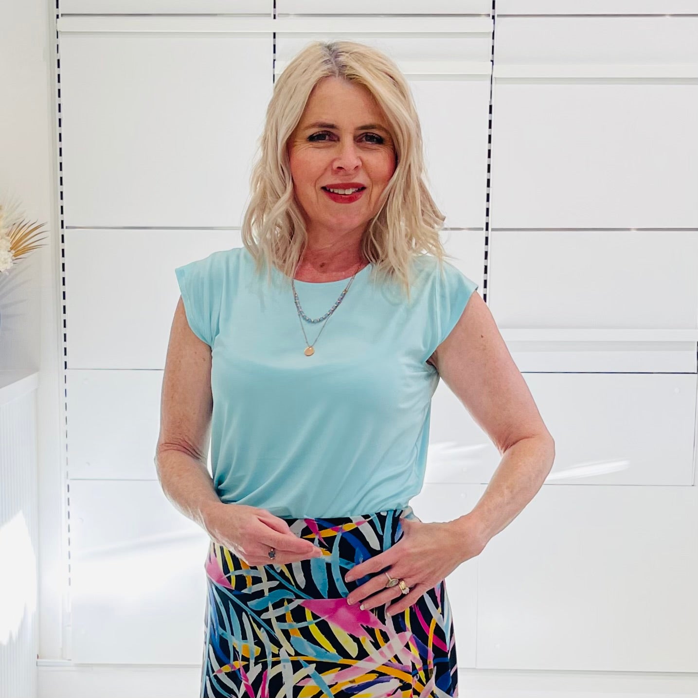 Woman wearing a light blue top and colorful skirt against a white tiled wall.