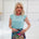 Woman wearing a light blue top and colorful skirt against a white tiled wall.
