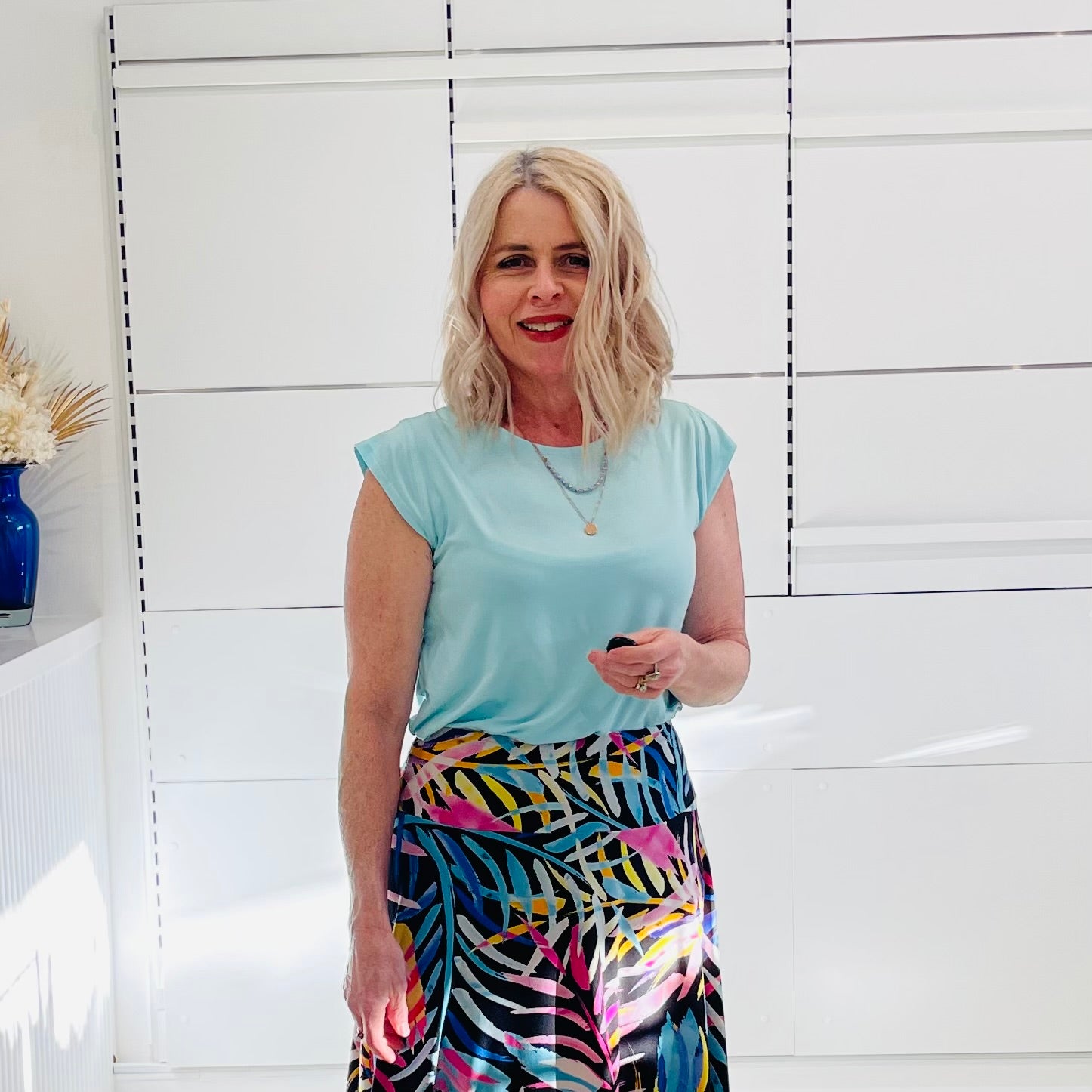 Woman wearing a light green  top and colorful skirt standing against a white wall.