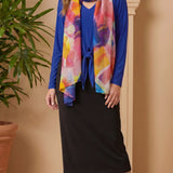 Woman wearing a blue cardigan, colorful scarf, black skirt, and brown boots against a beige wall.