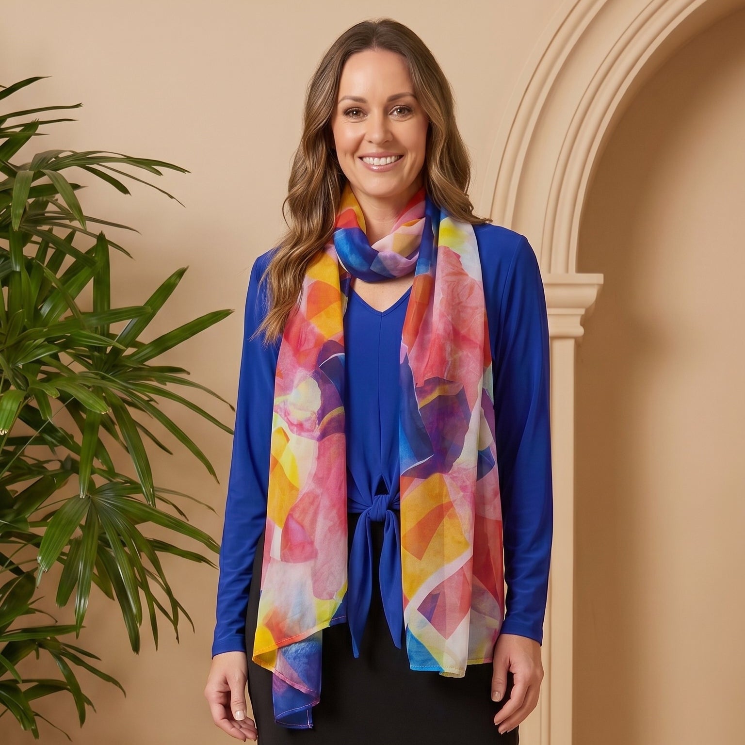 Woman wearing a blue top, black skirt, and tan boots with a colorful scarf in a room with a plant.