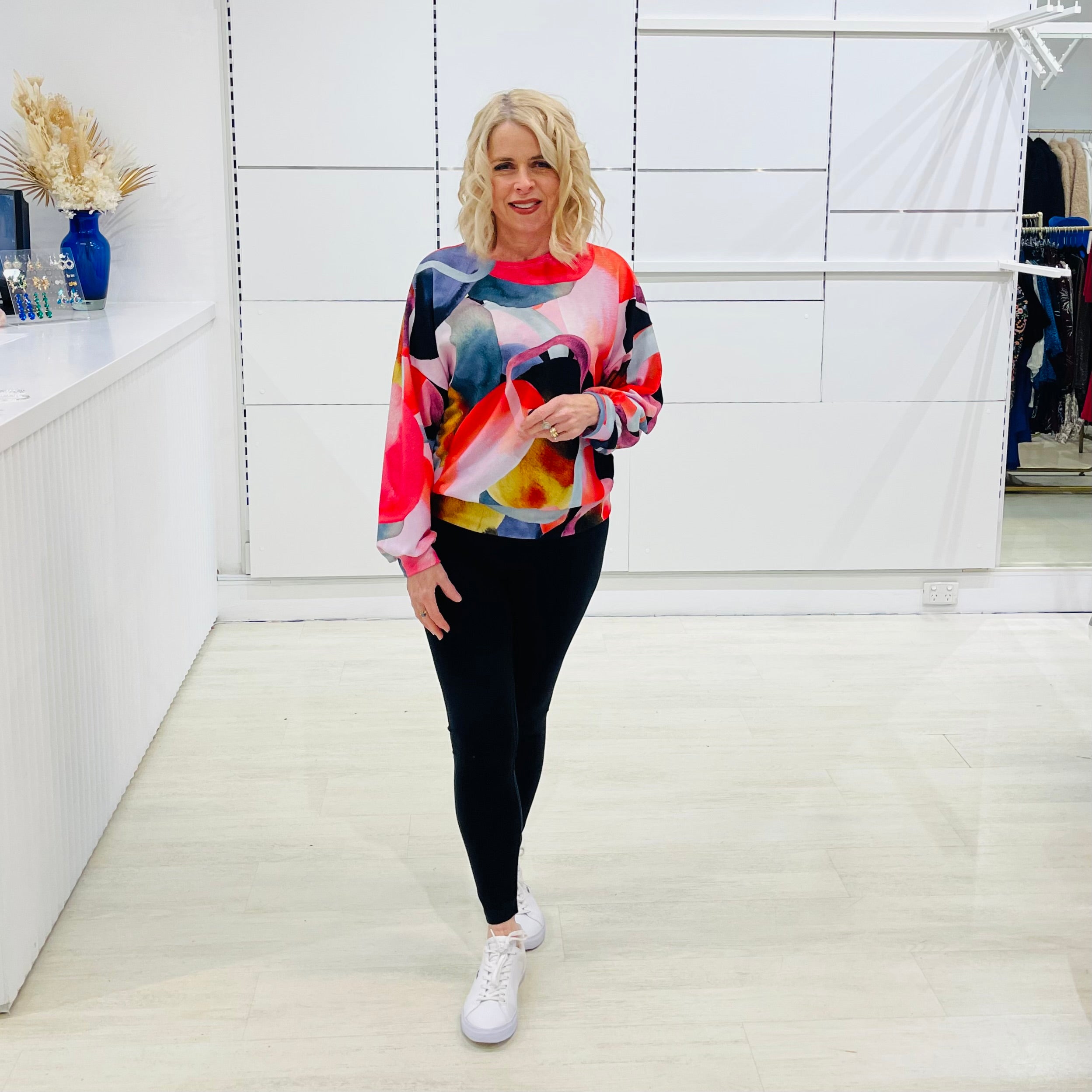 Woman wearing a colorful sweater and black leggings in a room with white walls and a shelf.