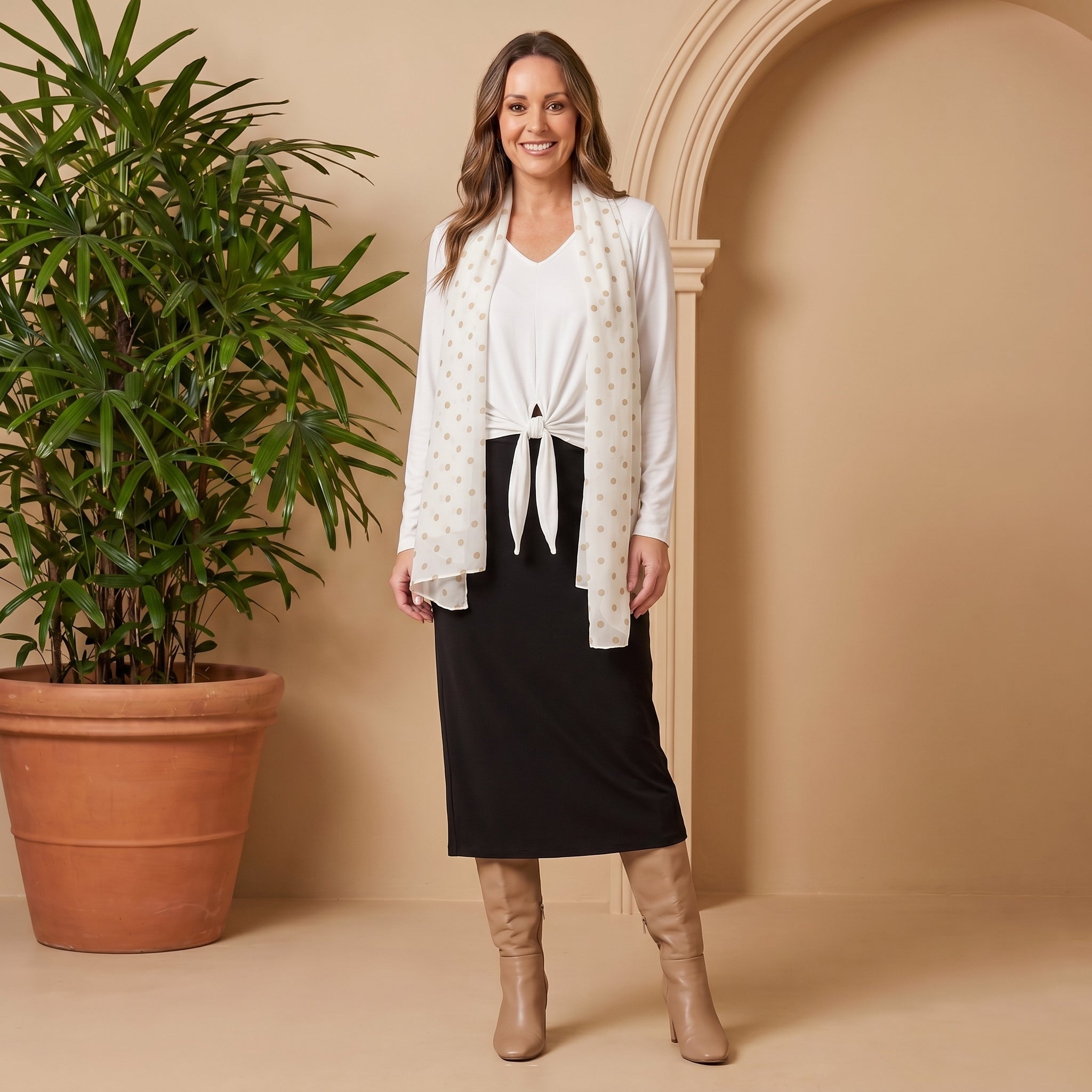 Woman in a white blouse and black skirt standing next to a potted plant against a beige wall.