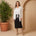 Woman in a white blouse and black skirt standing next to a potted plant against a beige wall.