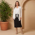 Woman in a white blouse and black skirt standing next to a potted plant against a beige wall.