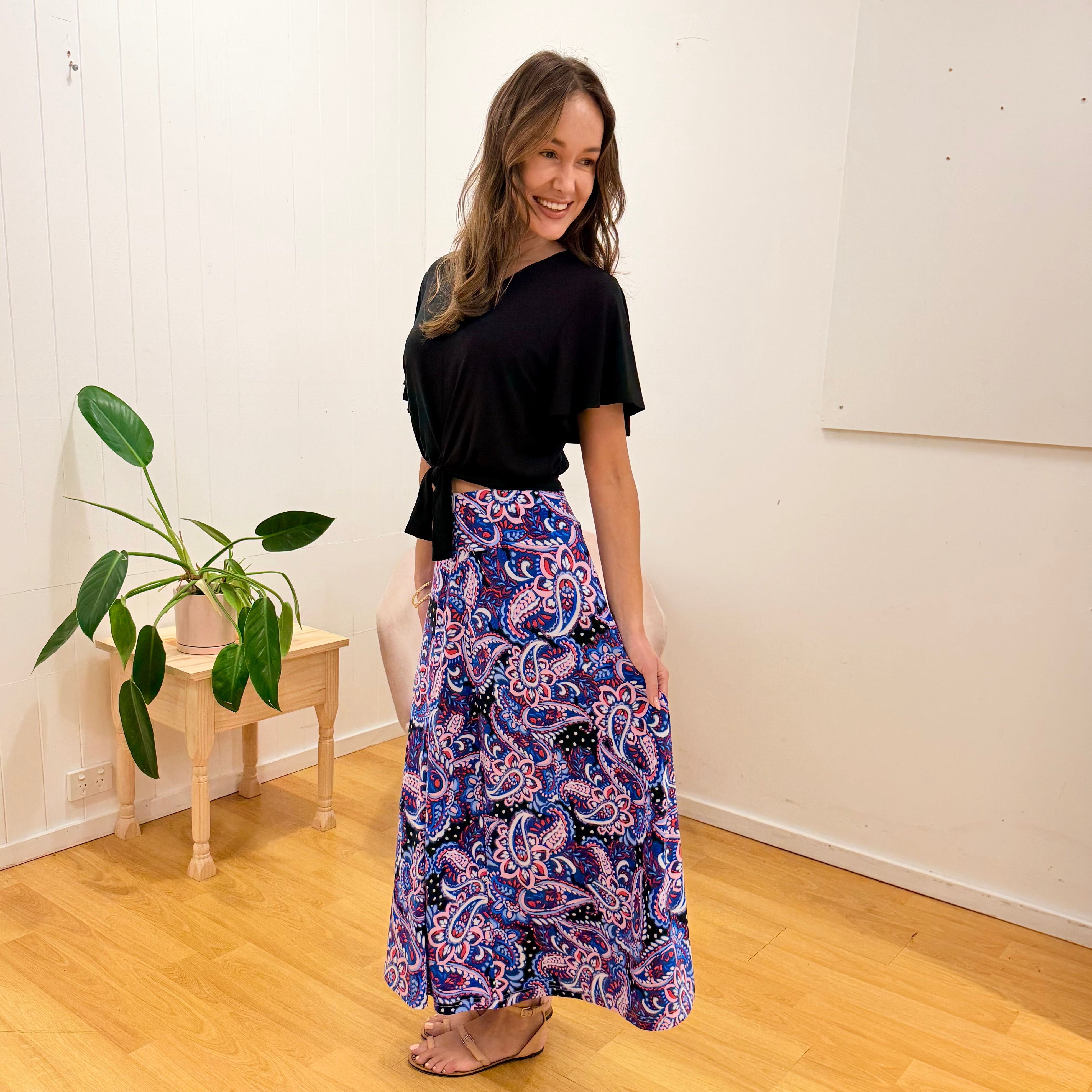 Woman wearing a black top and colorful paisley skirt standing in a room with a plant and wooden side table.