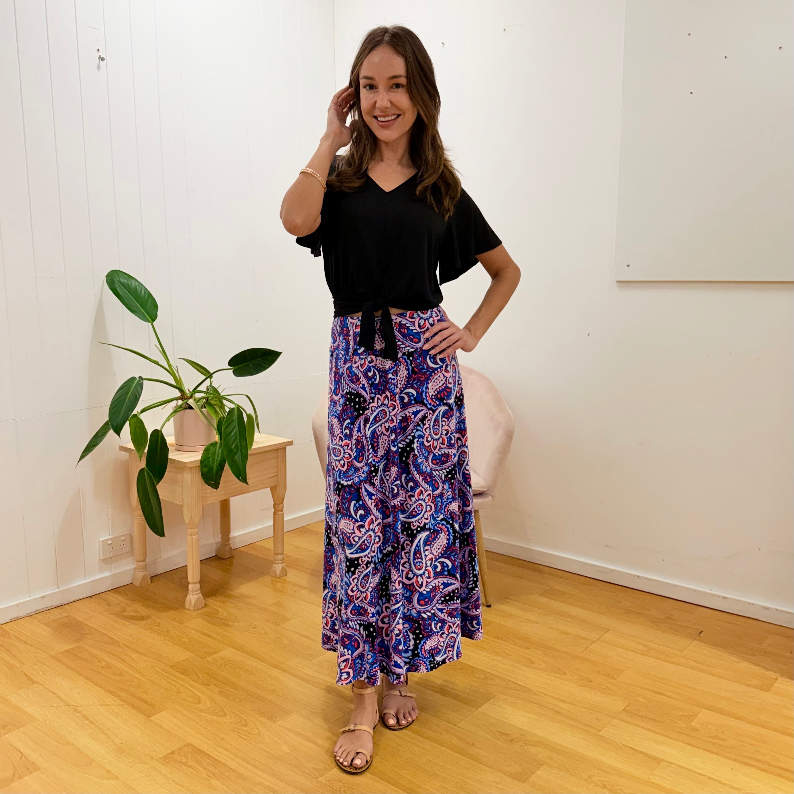 Woman wearing a black top and purple patterned skirt standing in a room with a plant and chair.