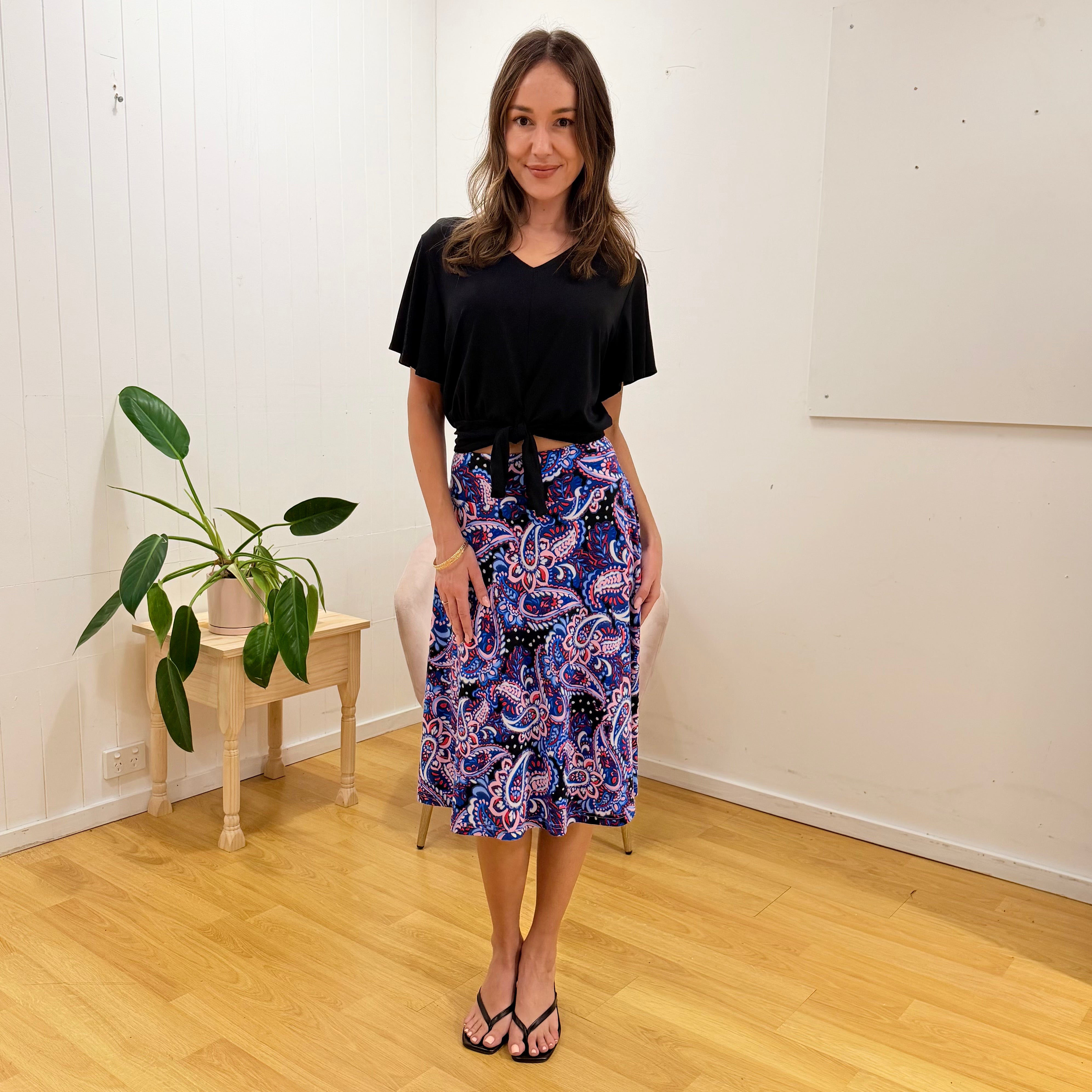 Woman wearing a black top and colorful skirt standing in a room with a plant and small table.