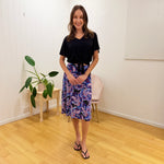Woman standing in a room with a plant and chair, wearing a black top and colorful skirt.