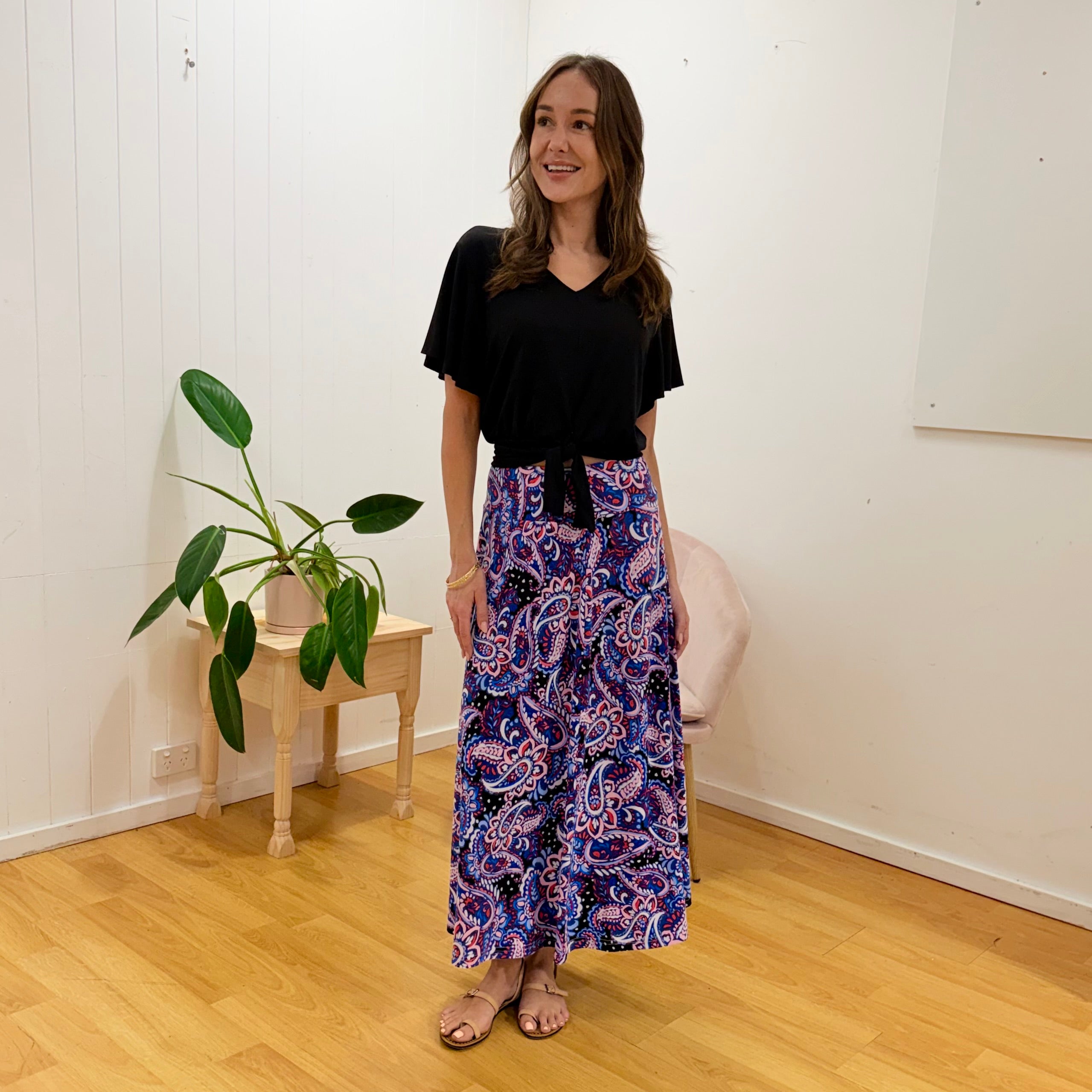 Woman wearing a black top and colorful patterned pants standing in a room with a plant and small table.