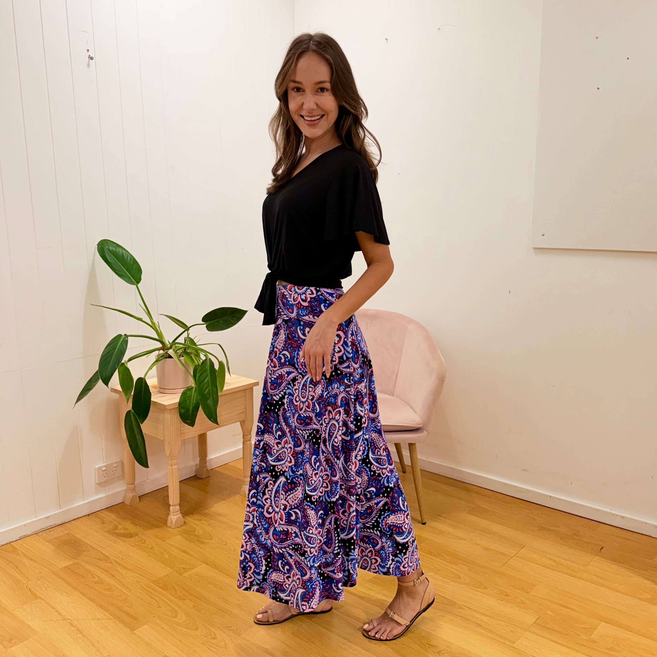 Woman wearing a black top and colorful patterned skirt standing in a room with a plant and chair.
