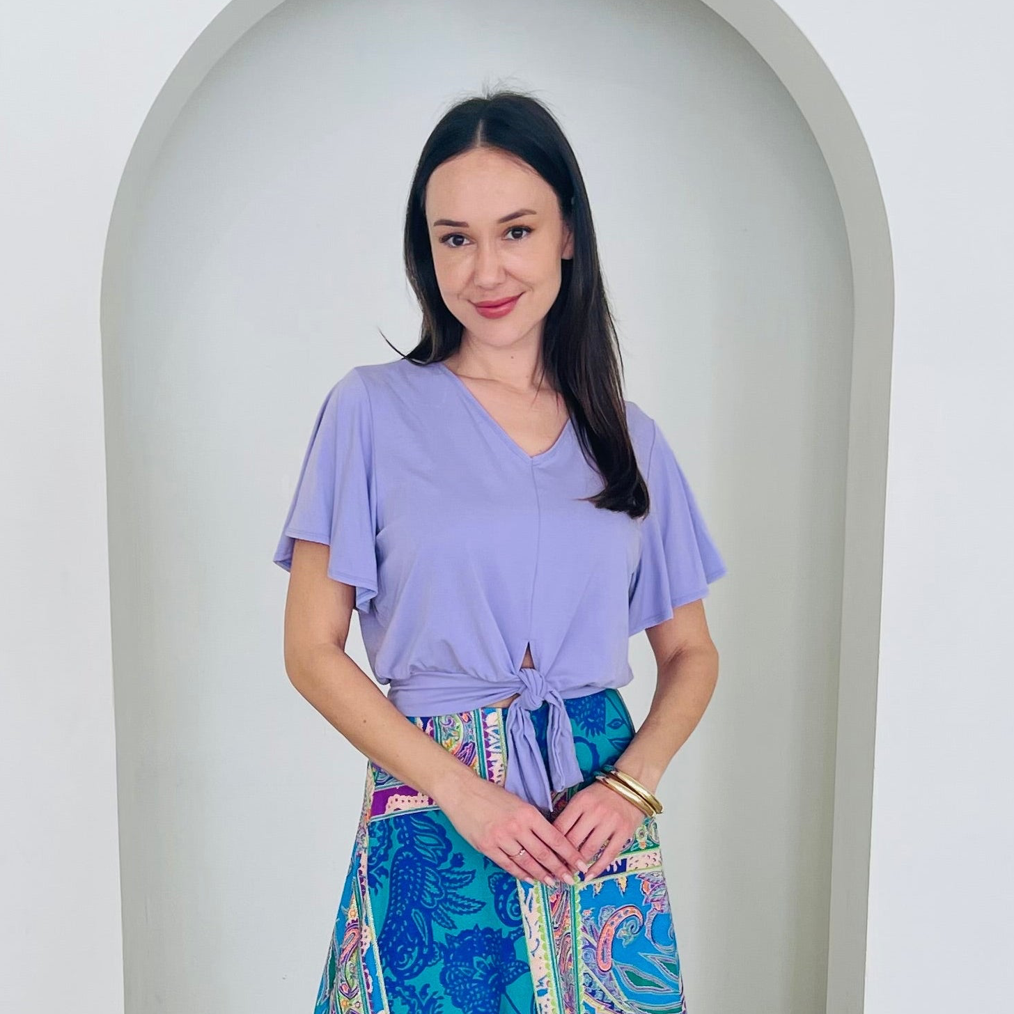Woman wearing a purple top and colorful skirt standing in front of a white archway.