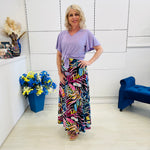 Woman in a colorful skirt and purple standing in a room with a blue chair and floral arrangement.