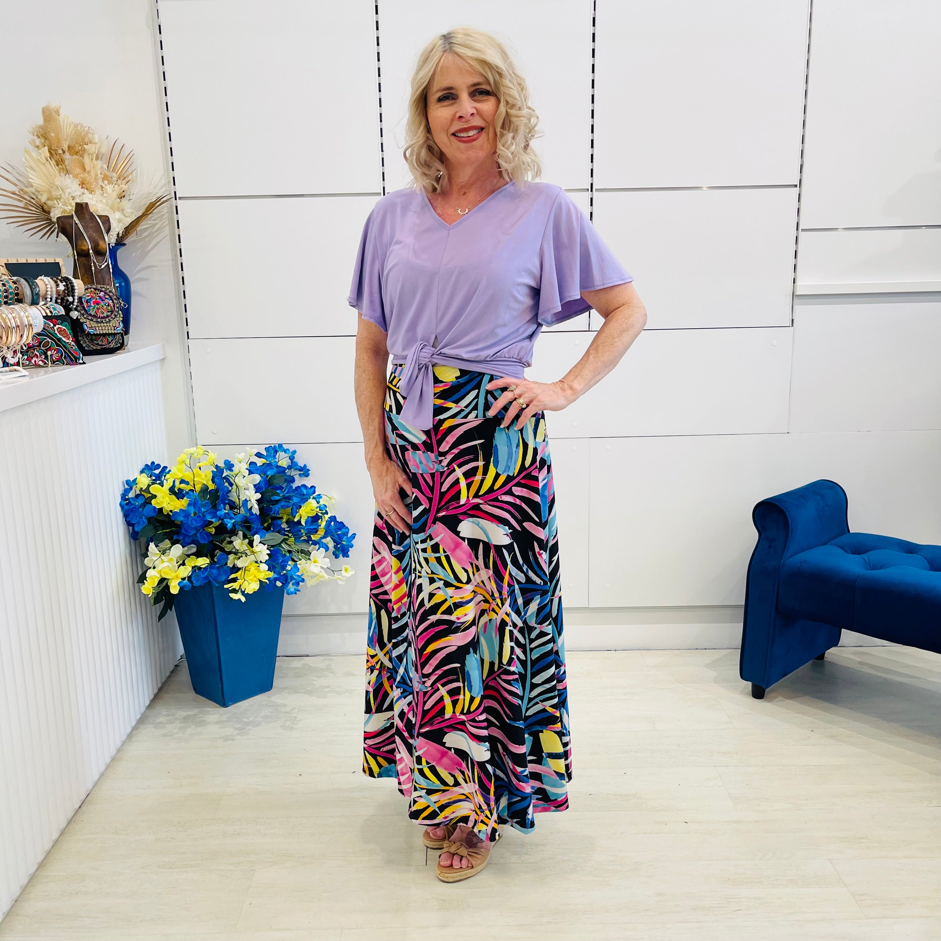 Woman in a colorful skirt and purple standing in a room with a blue chair and floral arrangement.