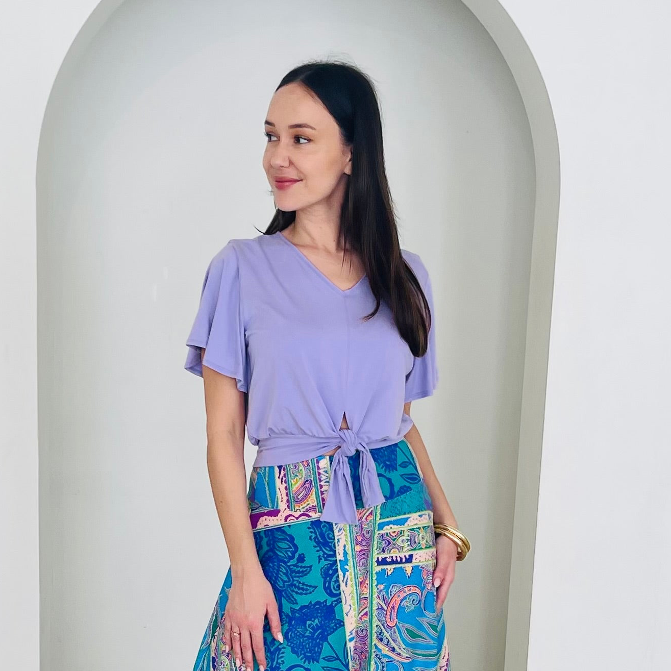 Woman wearing a purple top and a colorful skirt standing against a white archway.
