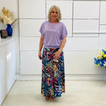 Woman wearing a light purple top and colorful patterned skirt standing in a room with white walls and a shelf.