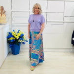 Woman wearing a purple top and colorful skirt standing next to a blue flower pot indoors.