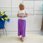 Woman in a white top and purple skirt standing in a room with a floral arrangement and a cabinet.