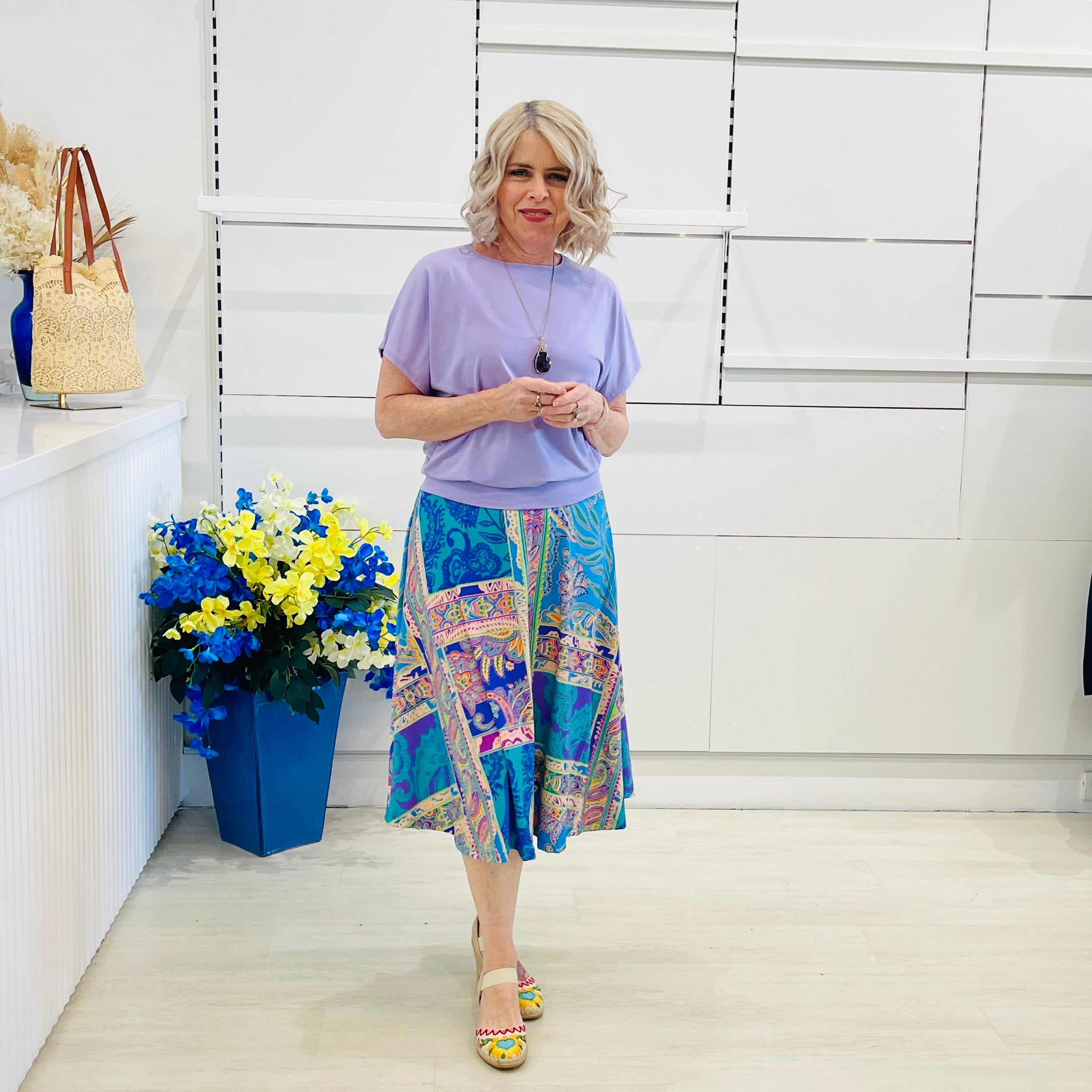 Woman wearing a purple top and colorful skirt standing next to a blue vase with flowers  against a white wall.