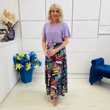 Woman wearing a purple top and colorful pants standing in a room with a blue chair and flower arrangement.