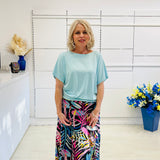 Woman wearing a light blue top and colorful skirt standing in a room with white walls and decorative elements.