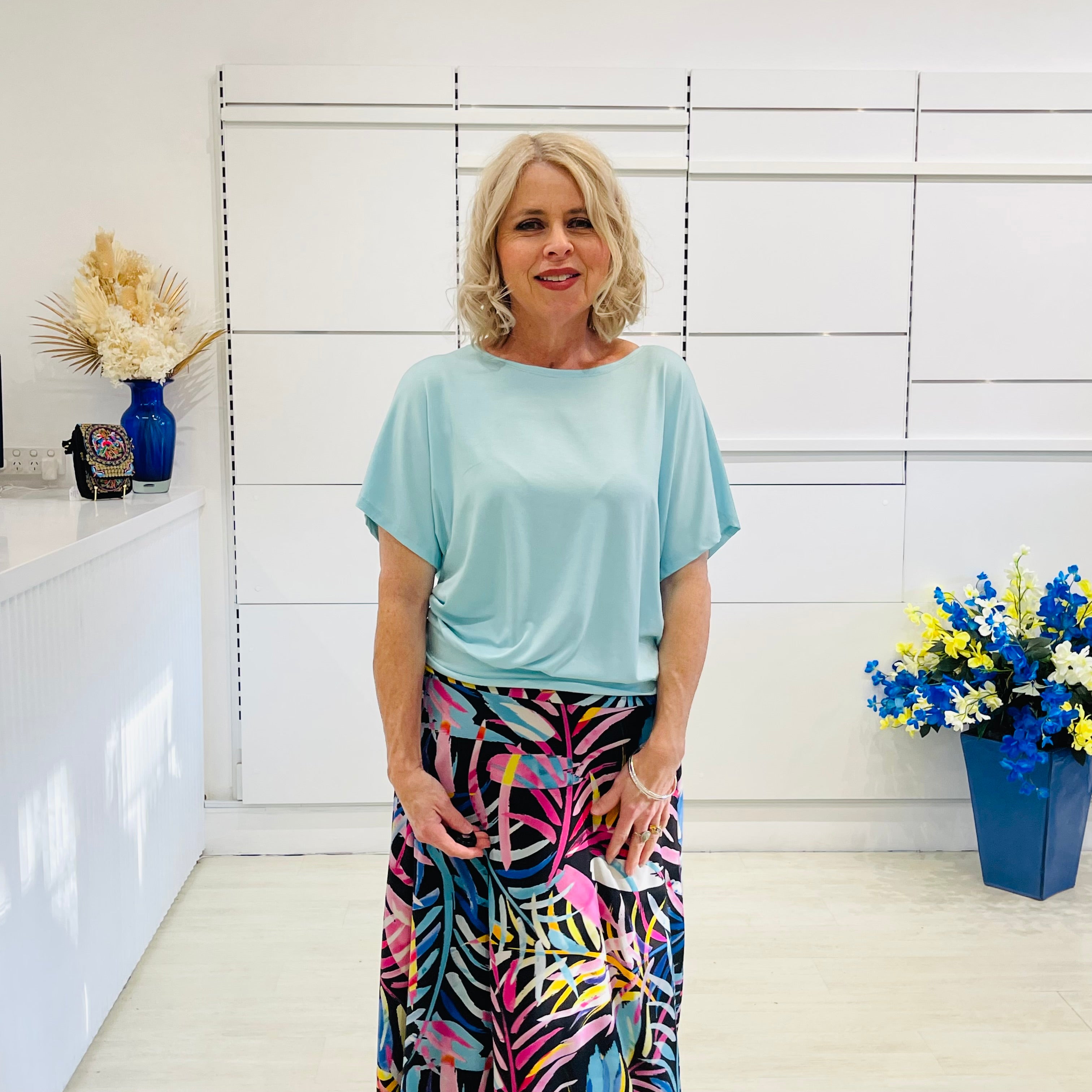 Woman wearing a light blue top and colorful skirt standing in a room with white walls and decorative elements.
