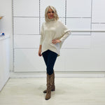 Cream knit, long sleeve, high neckline, asymmetrical hemline, winter 2025 jumper sweater. With black pants and and brown boots with embroidery.