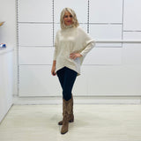 Cream knit, long sleeve, high neckline, asymmetrical hemline, winter 2025 jumper sweater. With black pants and and brown boots with embroidery.
