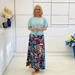 Woman wearing a light blue top and colorful floral skirt in a room with white walls and decorative elements.