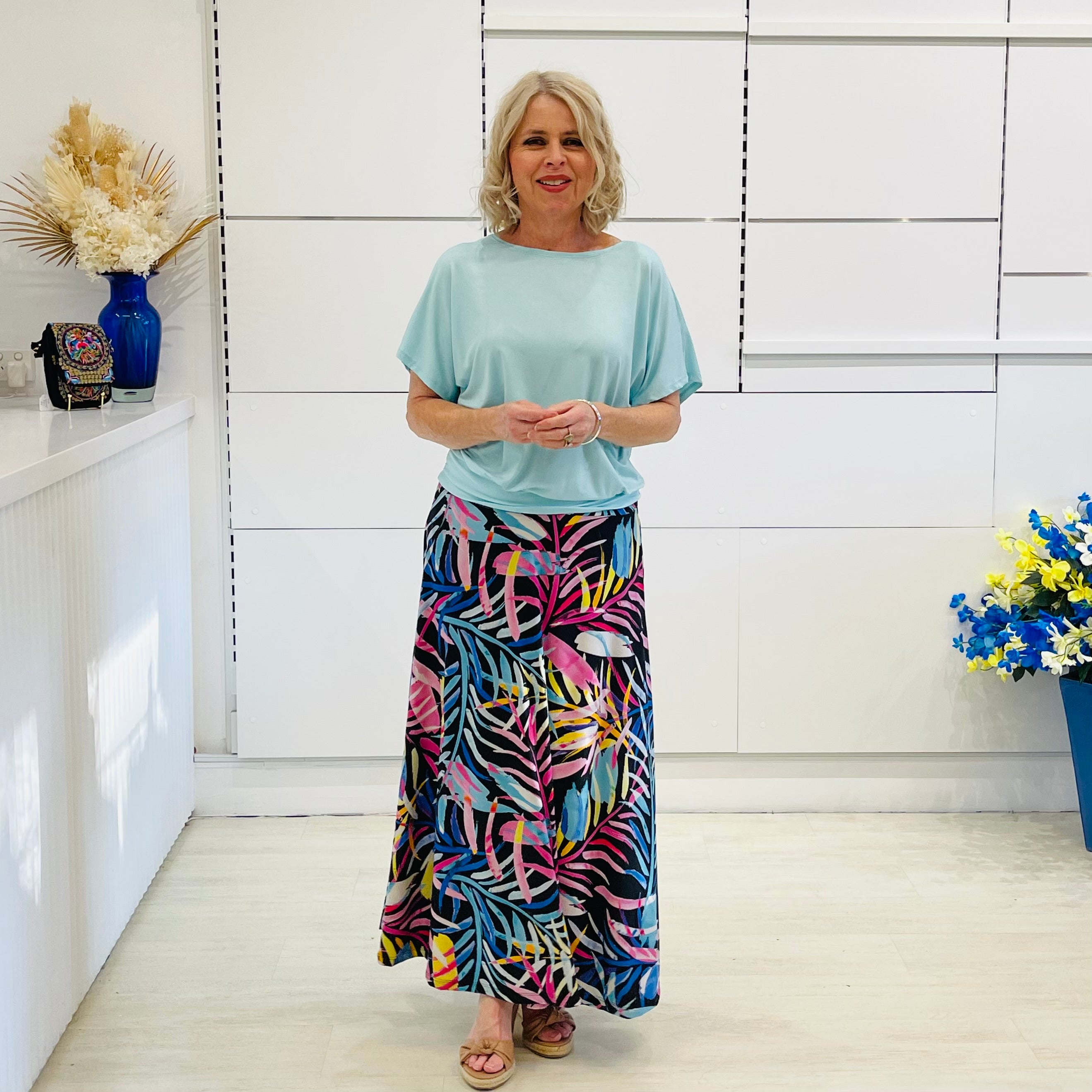 Woman wearing a light blue top and colorful floral skirt in a room with white walls and decorative elements.