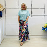 Woman wearing a light blue top and colorful skirt standing in a room with white walls and a wooden floor.