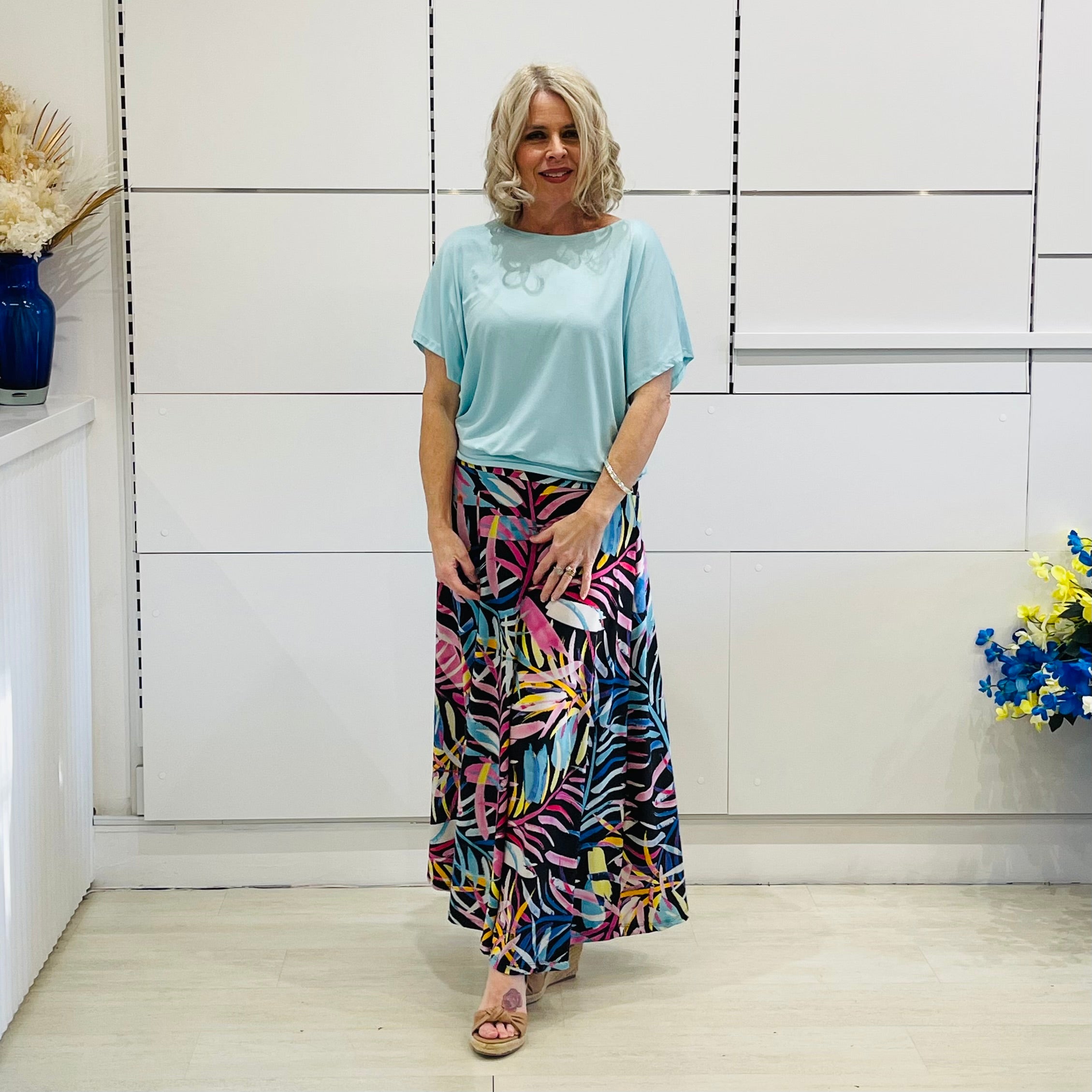 Woman wearing a light blue top and colorful skirt standing in a room with white walls and a wooden floor.