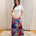A woman stands indoors wearing a white short-sleeve top and a long, flowing maxi skirt with a vivid abstract watercolor print in red, blue, purple, and yellow. She is posed on a patterned rug in front of white curtains.