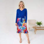 Woman wearing a blue top and colorful skirt in a room with a chair and plant.