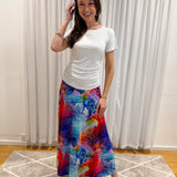 A woman modeling a white short-sleeve top and a long, colorful abstract-print skirt in front of white curtains and a beige rug.