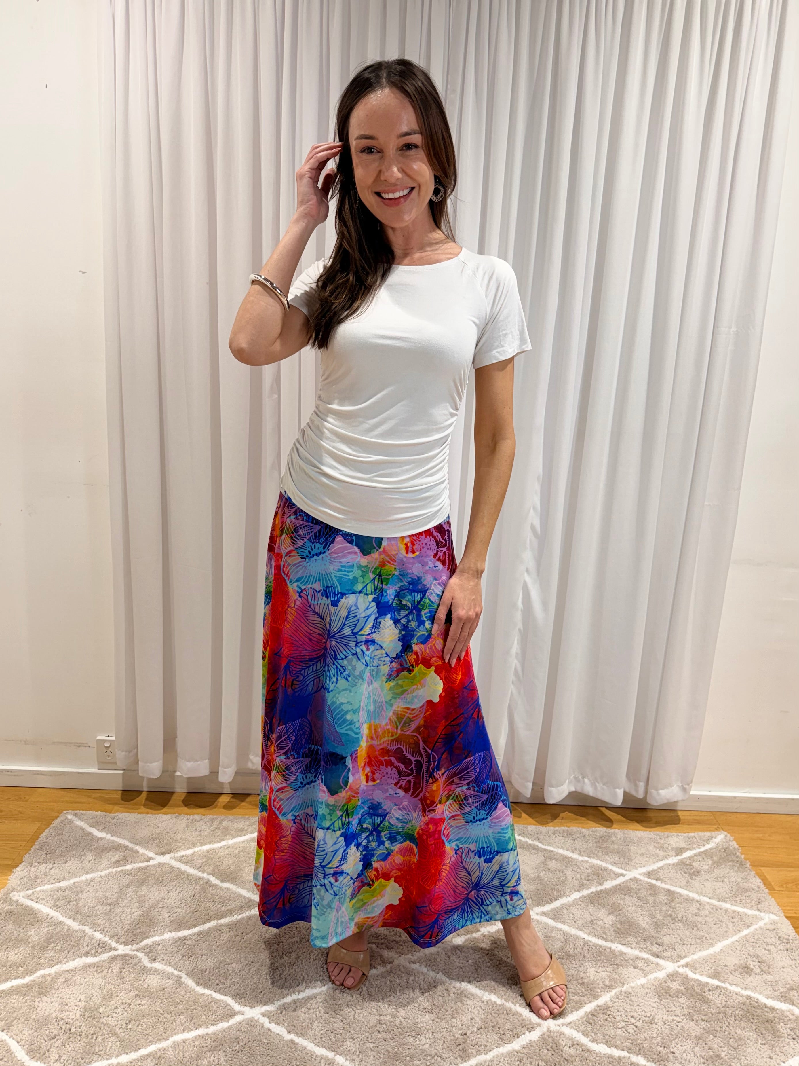 A woman modeling a white short-sleeve top and a long, colorful abstract-print skirt in front of white curtains and a beige rug.