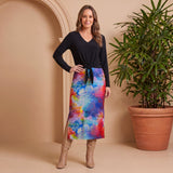 Woman wearing a black top and colorful skirt standing next to a potted plant against a beige wall.