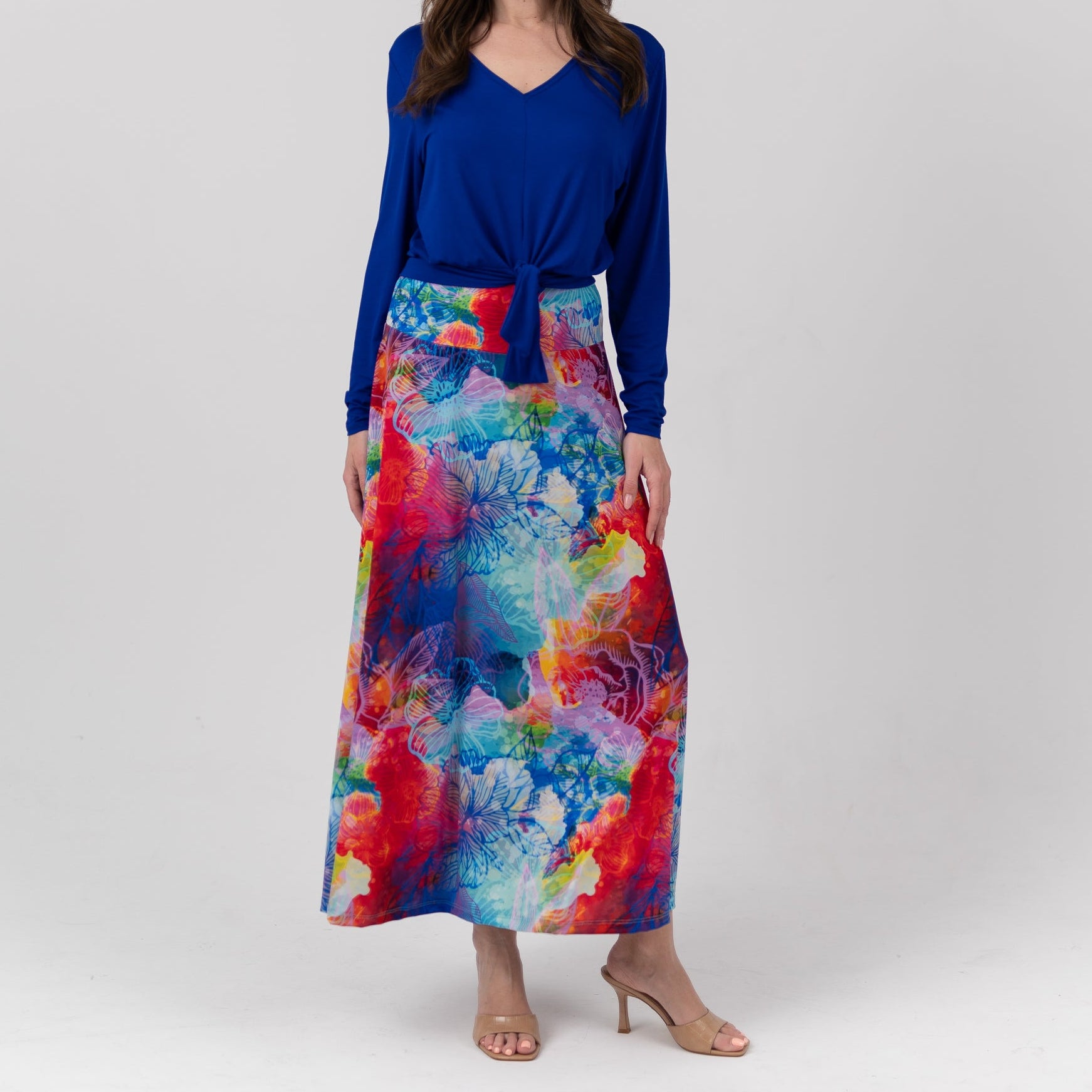 A woman modeling a long-sleeve wrap-style dress with a vibrant blue V-neck top and a multicolor abstract floral print skirt, standing against a light gray studio background.